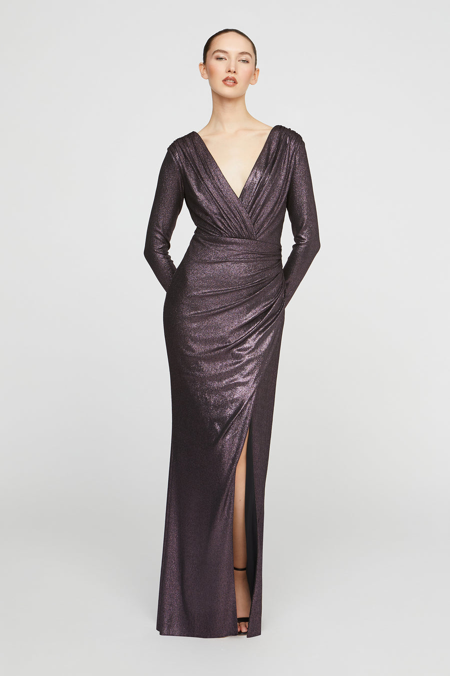 Woman wearing a long, dark purple evening gown with a deep V-neck and slit on a light gray background